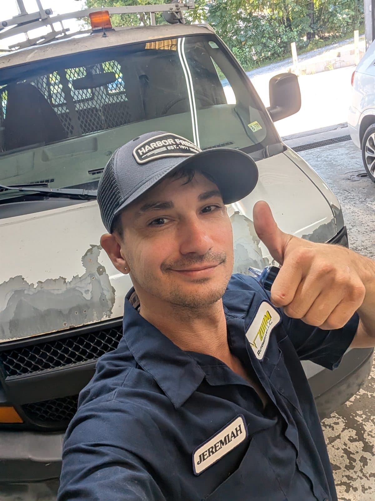 Jeremiah Gilpin, owner of Zoom Automotive at 2014 12th Street in Lynchburg, Virginia, in a navy Zoom Automotive work shirt with the shop logo and his name patch, standing at the open bay door