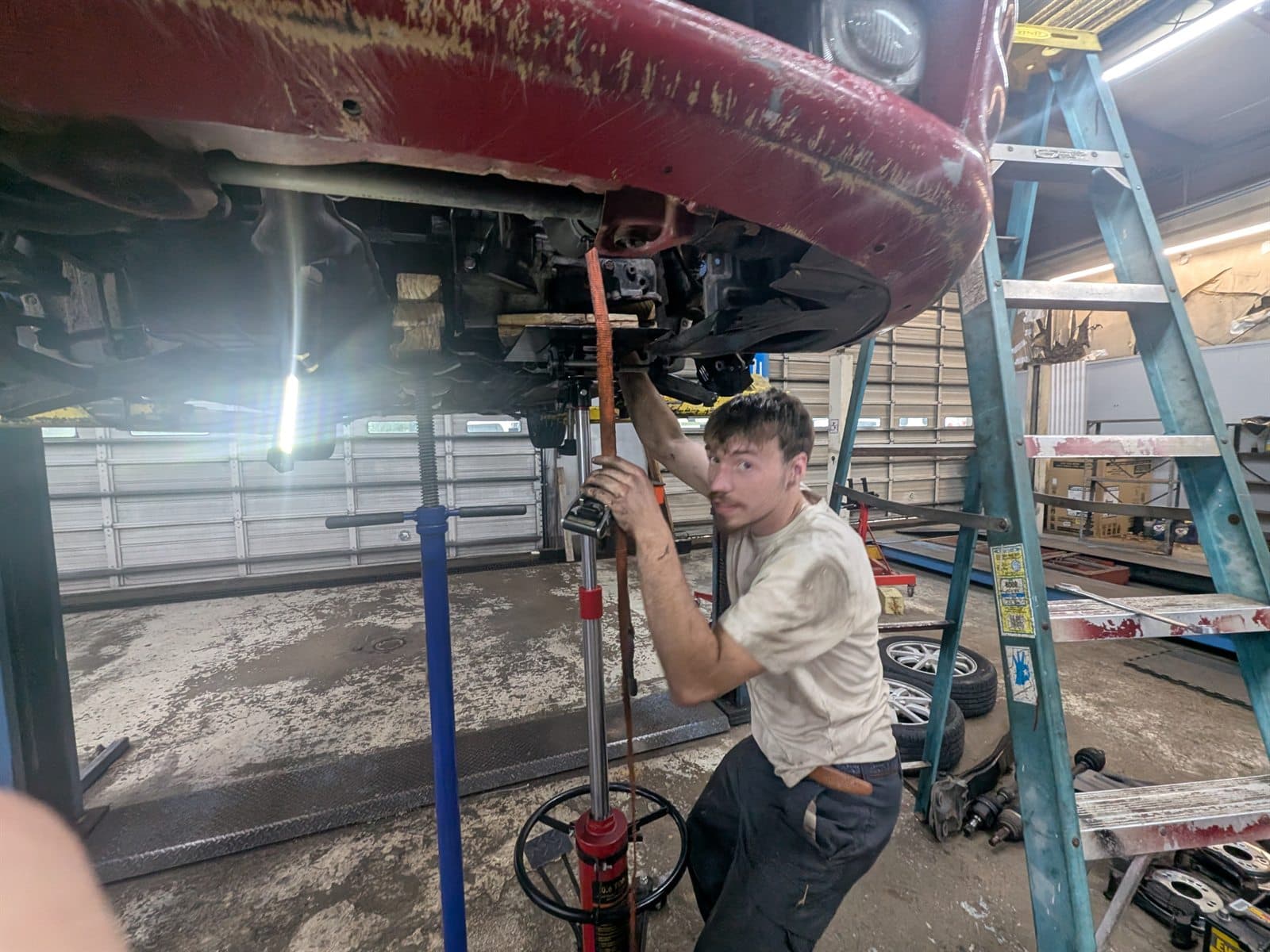 A technician at Zoom Automotive in Lynchburg, Virginia, steadying a transmission jack under a red vehicle raised on a two-post lift in the service bay